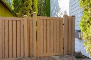 A tan vertical-plank fence with a central gate standing beside a white house.