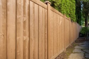 A tall, light brown vertical fence with a wood-grain texture runs along a path bordered by a hedge.