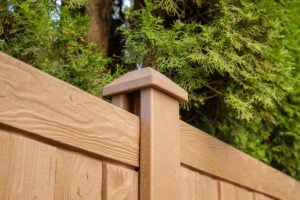 Brown composite fence post with a flat cap, positioned in front of green evergreen foliage.