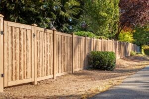 A tall, light-brown wood-look fence runs along a paved path next to trees and shrubs.