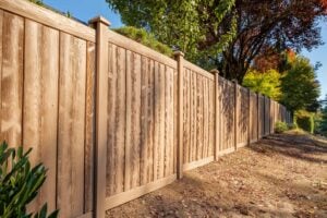 A long, tan privacy fence with a wood-grain texture lines a yard with trees and dry ground.