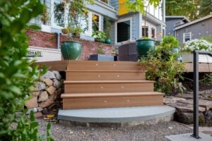 Three wooden steps lead up to a deck with large potted plants and a brick foundation.