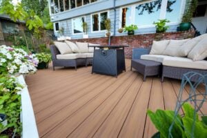 A patio deck featuring wicker outdoor furniture with beige cushions and a central fire pit table in front of a house.