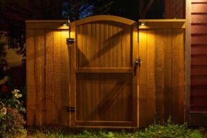 A wooden fence with a centered arched gate, illuminated at night by two small lights mounted on the fence posts.