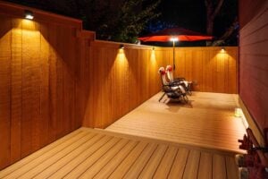 A wooden deck at night, lit by wall-mounted lights, featuring two reclining chairs under a red patio umbrella.