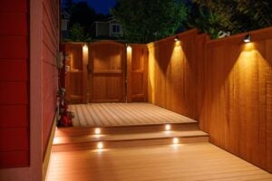 Wooden deck with two illuminated steps and wall-mounted exterior lights illuminating a wooden fence and gate at night.