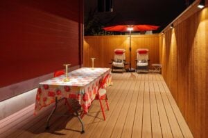 An outdoor deck at night with a table set for two, two reclining chairs under a red umbrella, and warm wall lighting.