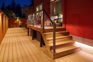 A tiered wooden deck with metal railings and illuminated stairs leading up to a sliding glass door against a red house wall.