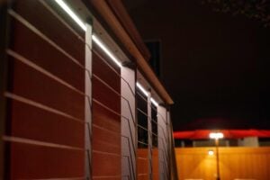 A deck railing with integrated LED strip lighting at night, with a blurred red umbrella and yard light in the background.