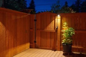 A wooden gate and fence section on a deck at night, illuminated by a small light above the gate next to a potted plant.