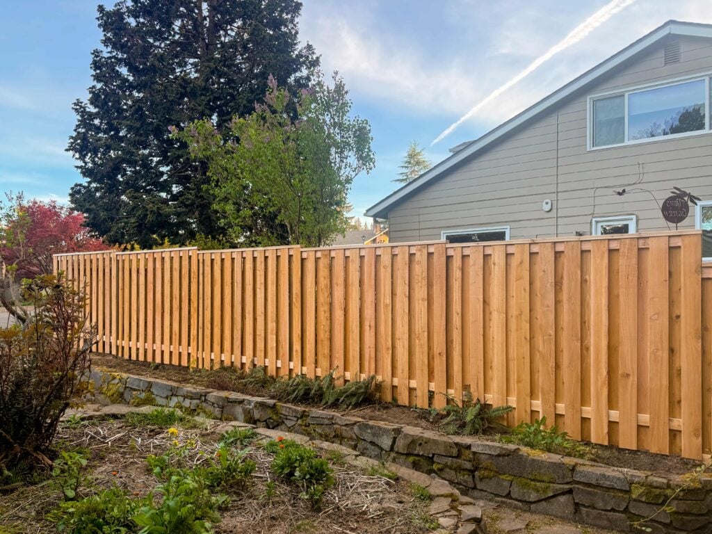 A new wooden fence runs along a stone retaining wall in a backyard, with plants and trees nearby and a house in the background under a blue sky with clouds.