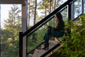 A woman sits on a wooden staircase and reads a book, with a forest view visible through large glass windows.