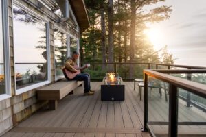 A man plays an acoustic guitar while sitting on a wooden bench on a deck next to a lit fire pit.