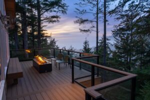 A wooden deck with two chairs and a rectangular fire pit overlooks a forest and a distant, misty sunset.