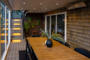 A wooden outdoor dining table with a vase of greenery sits on a deck beside a set of stairs with illuminated steps and a glass sliding door.