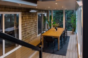 A wooden outdoor dining table with black chairs on a deck, situated inside a screened-in porch with wood-shingled walls.