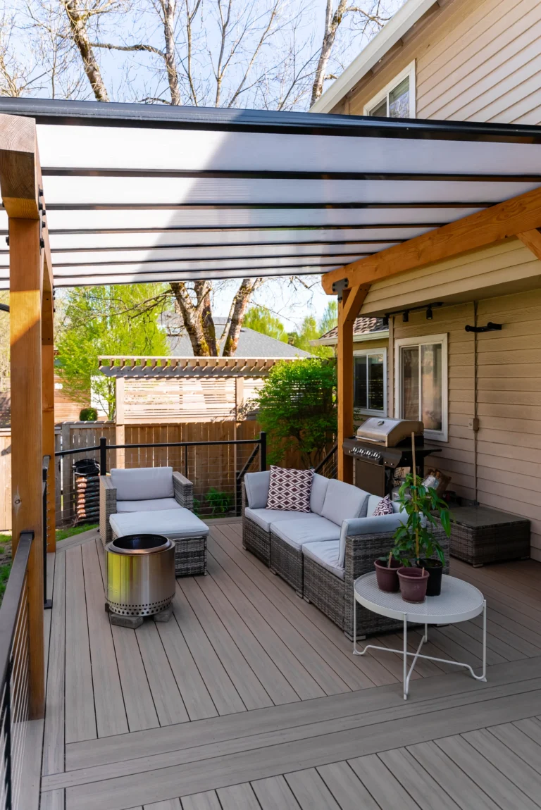 Modern backyard patio with a pergola, wicker outdoor sofa and chairs with white cushions, a round fire pit, potted plants on a small table, and a grill, adjacent to a beige house on a sunny day.