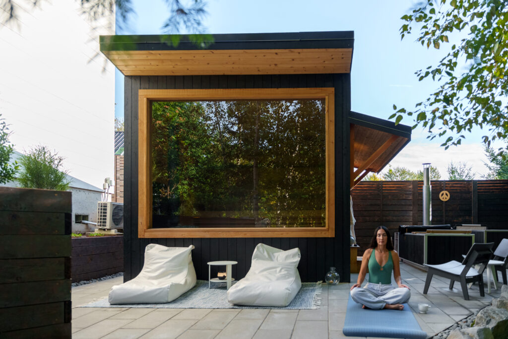 A woman practices yoga on a mat in a modern patio with two white lounge chairs, a small table, and a black shed with a large window and wooden trim in the background.