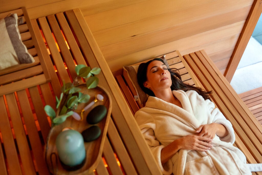 A woman in a white robe relaxes on a wooden bench in a spa or sauna, eyes closed. A wooden tray with a candle, stones, and greenery is nearby, creating a peaceful atmosphere.