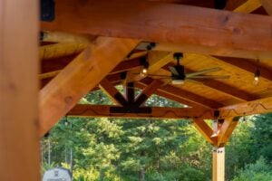 A timber-frame pavilion structure with a ceiling fan and string lights, overlooking a wooded area.
