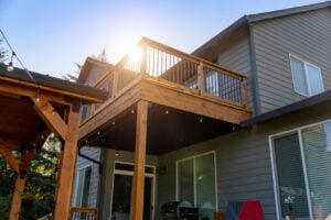 A two-story house with a wooden deck and string lights, viewed from the patio below as the sun shines over the railing.