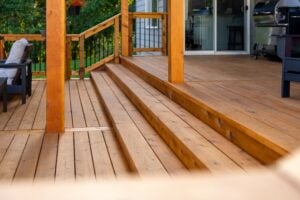 A wooden deck with a set of three wide steps leading up to a sliding glass door and a grill.