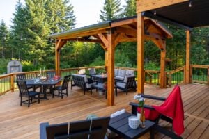 A wooden deck featuring a dining area with a table and chairs, and a covered lounge area with outdoor sofas and a fire table, surrounded by a forest.