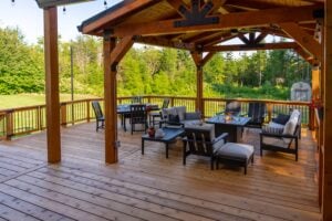 A covered wooden deck furnished with a dining table, a lounge area with a fire pit, and armchairs, overlooking a grassy yard and forest.