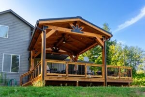 A wooden deck with a large covered pavilion featuring a peaked roof, ceiling fans, and string lights, attached to a gray house.