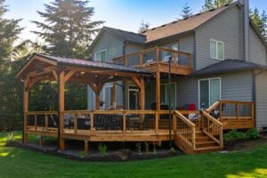 A two-story grey house with a large, multi-level wooden deck featuring a covered area with string lights and stairs leading to the yard.