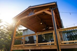 A wooden patio gazebo with a ceiling fan, string lights, and railings overlooking a backyard at sunset.