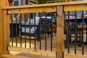 Wooden deck railing with vertical black metal balusters overlooking a patio seating area.