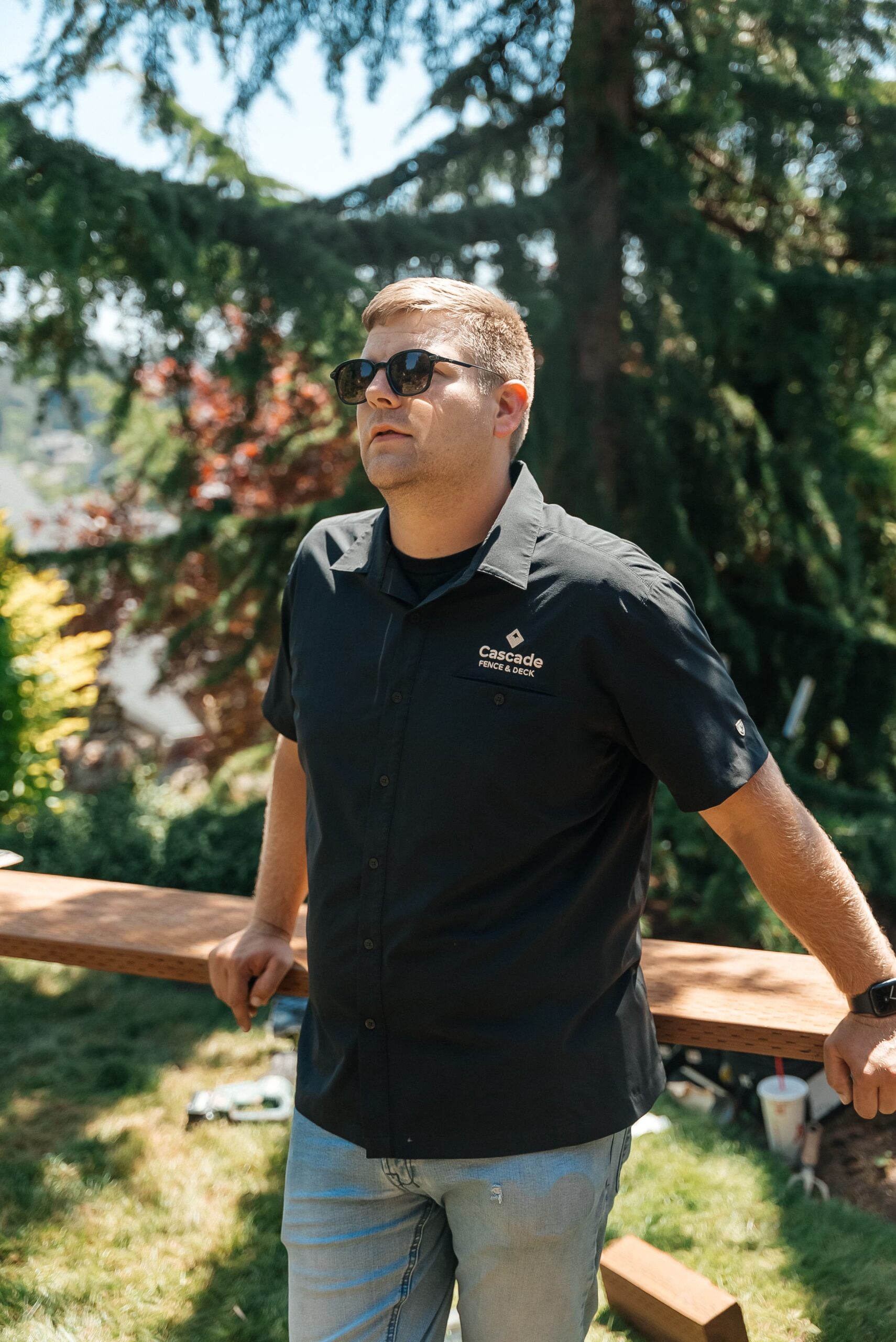 A man wearing sunglasses and a black Cascade Fence & Deck shirt stands outdoors on grass, leaning against a wooden beam, with trees and greenery in the background.