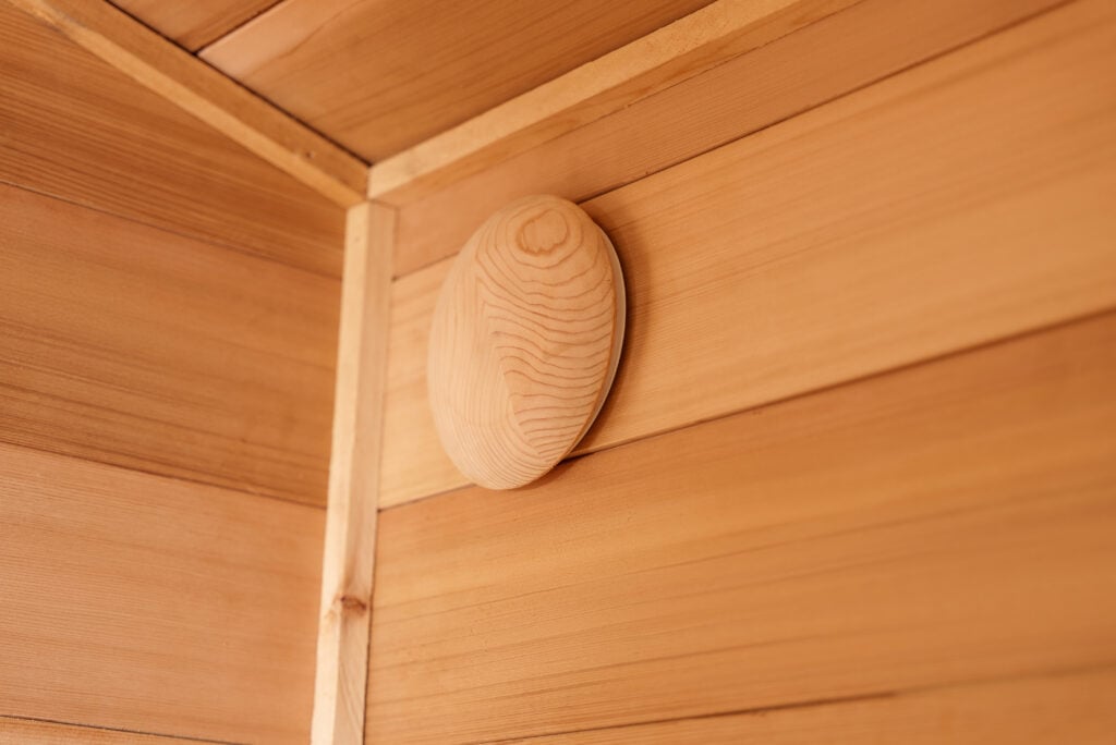 A close-up of a wooden vent cover on a wall made of horizontal wooden panels, positioned in the corner where two walls and the ceiling meet.
