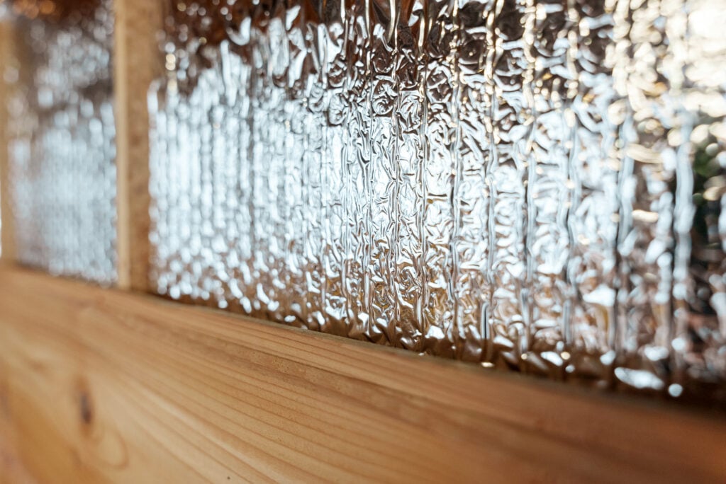 Close-up of a wooden surface with a sheet of crinkled reflective foil insulation behind it, showing texture and light reflections.