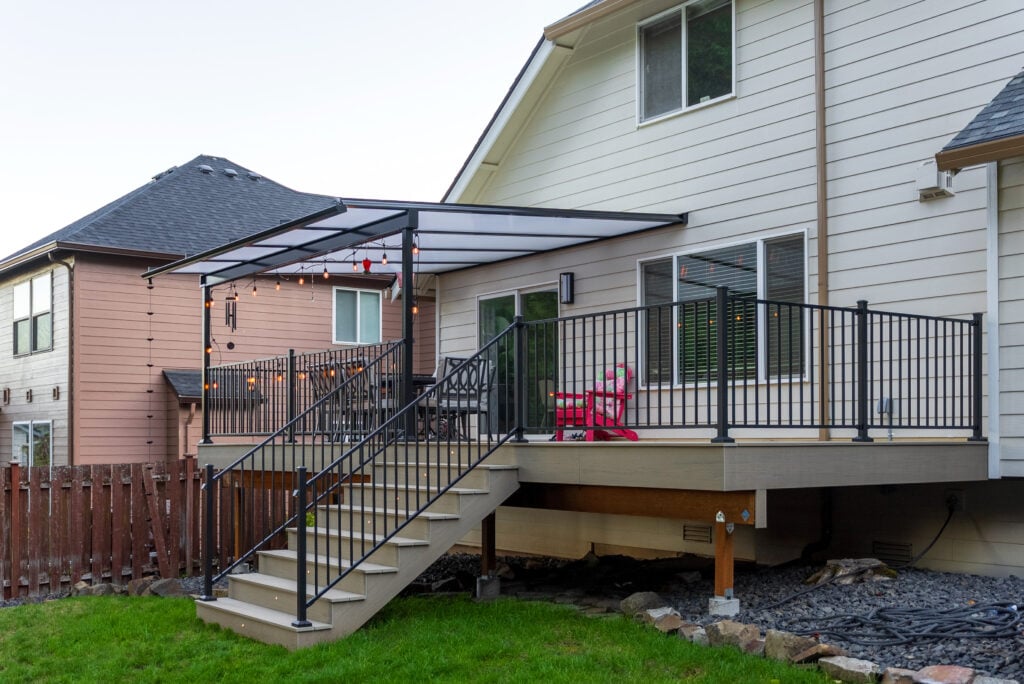 A raised backyard deck with black railings, string lights, a canopy, and two pink chairs. Stairs lead down to a green lawn. Two houses and a wooden fence are visible nearby.
