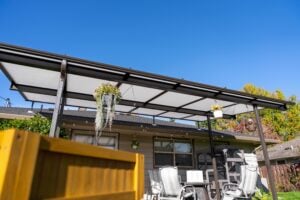 A patio covered by a dark-framed translucent roof with hanging planters, featuring outdoor furniture against the side of a house.