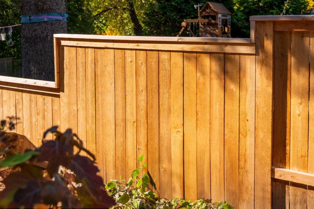 A tall wooden privacy fence stands in a sunlit backyard with some greenery in the foreground and a play structure partly visible in the background.