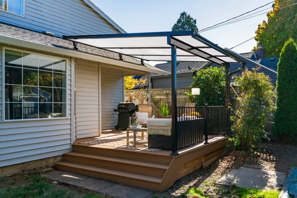 A backyard patio with a covered pergola, outdoor seating, a grill, plants, and a small table. The patio is connected to a beige house and accessed by wooden steps.