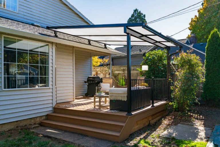 A backyard patio with a modern pergola, outdoor seating, a grill, and stairs leading to a grassy yard beside a beige house. Shrubs and a wooden fence are in the background.