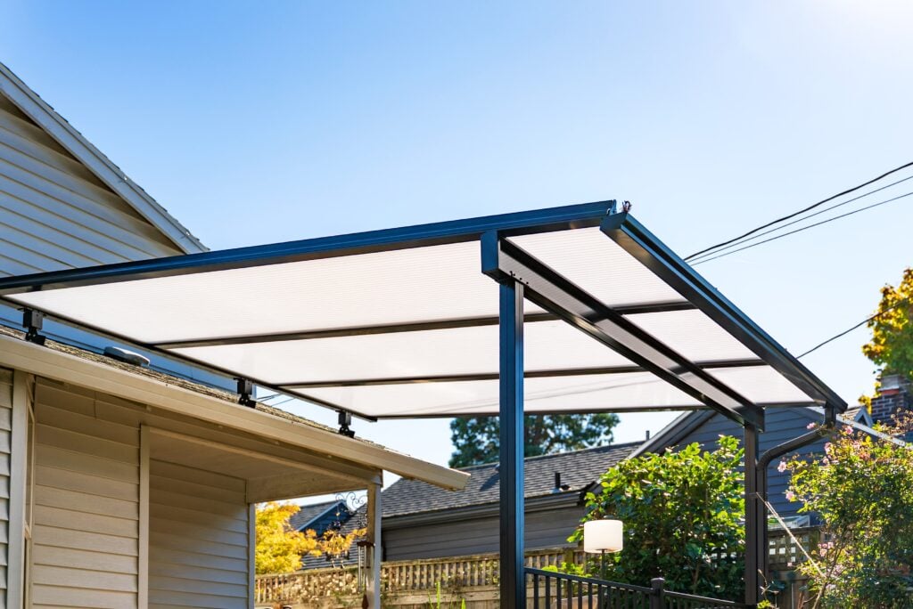 A modern, black metal patio cover with a translucent roof is attached to a house, providing shade in a backyard under a clear blue sky.
