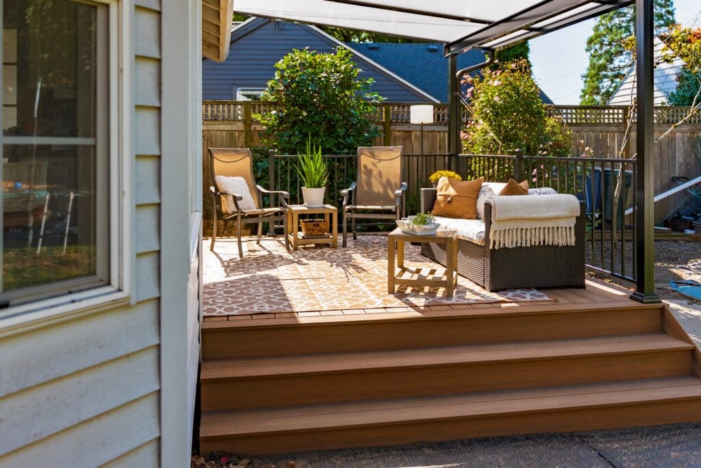 A cozy outdoor patio with chairs, a small table, a wicker sofa with a blanket, and a patterned rug under a pergola, surrounded by greenery and wooden fencing.