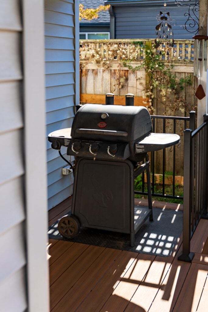 A black gas grill with three knobs sits on a small wooden deck, surrounded by a black metal railing, beside a blue house. There is a wooden fence and greenery in the background.