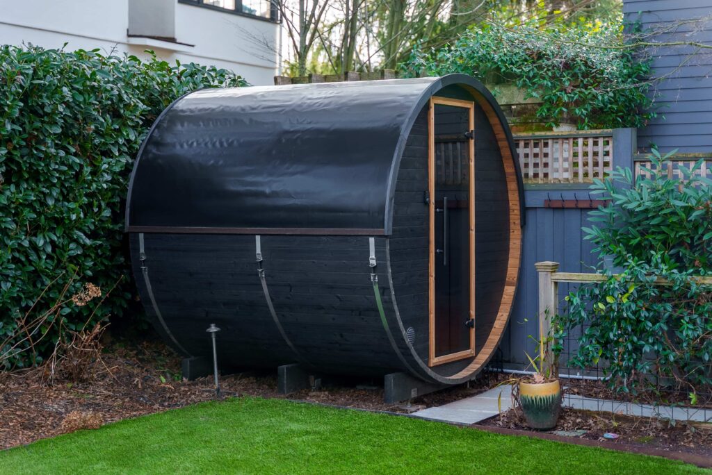 A barrel-shaped black sauna with a wooden door sits in a garden, surrounded by green bushes and a manicured lawn, next to a modern house with blue siding.