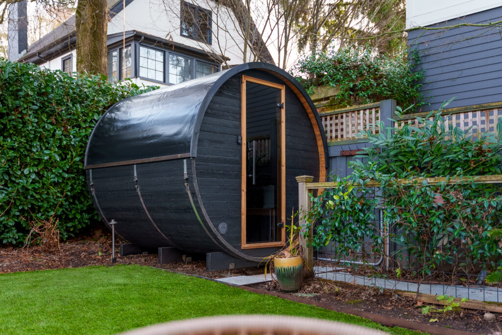 A black, barrel-shaped outdoor sauna sits on a raised platform in a garden, surrounded by green shrubs and trees, with a modern house partially visible in the background.