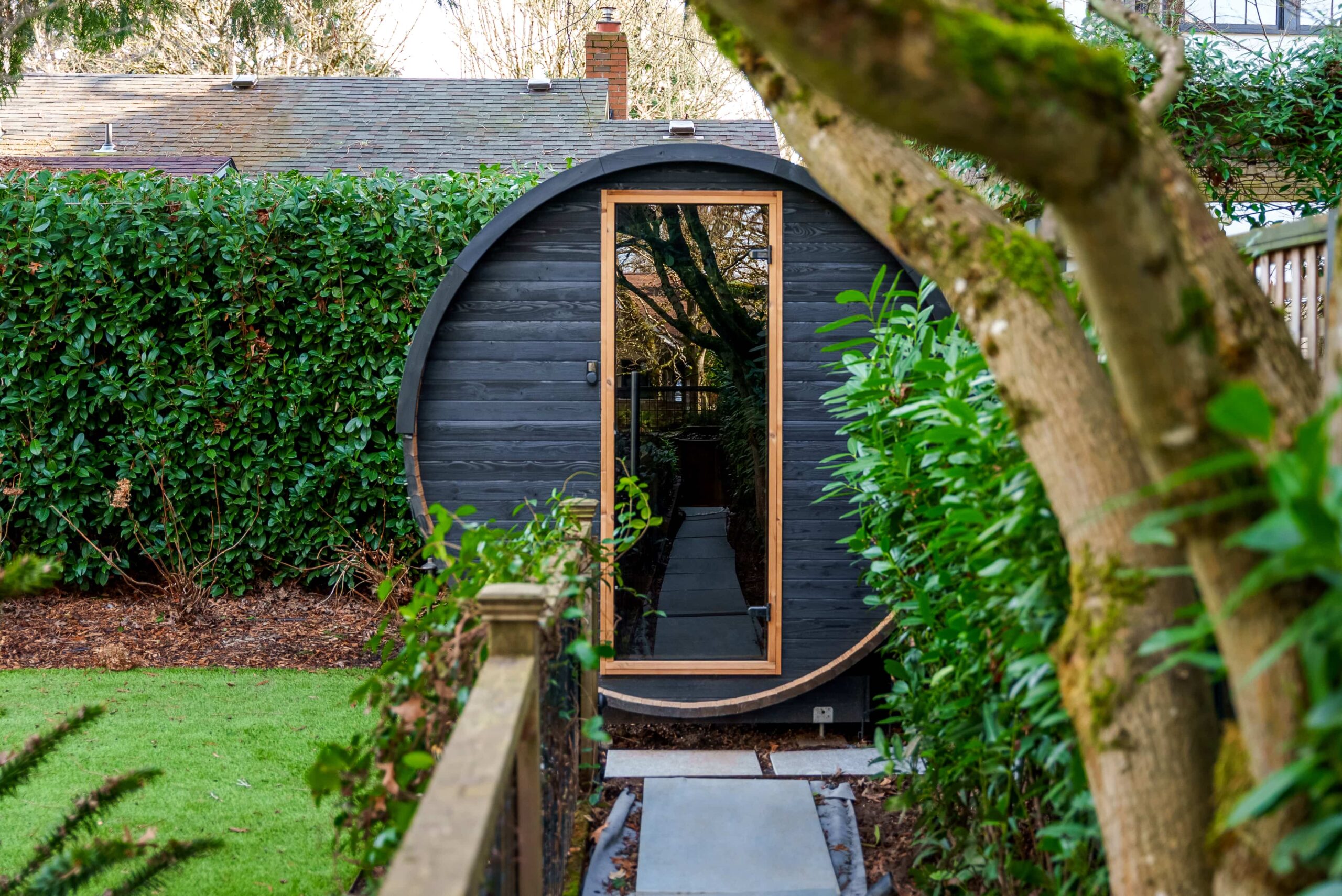 A modern, black, barrel-shaped garden shed with a tall glass door, surrounded by lush green hedges and trees, sits at the end of a stone path in a landscaped backyard.