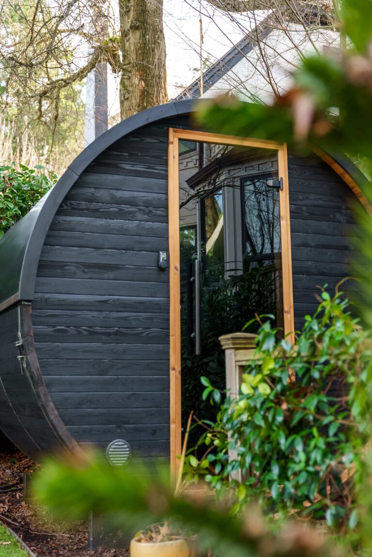 A black, barrel-shaped wooden cabin with a glass door sits among green bushes and trees, partially obscured by branches, in a wooded garden setting.