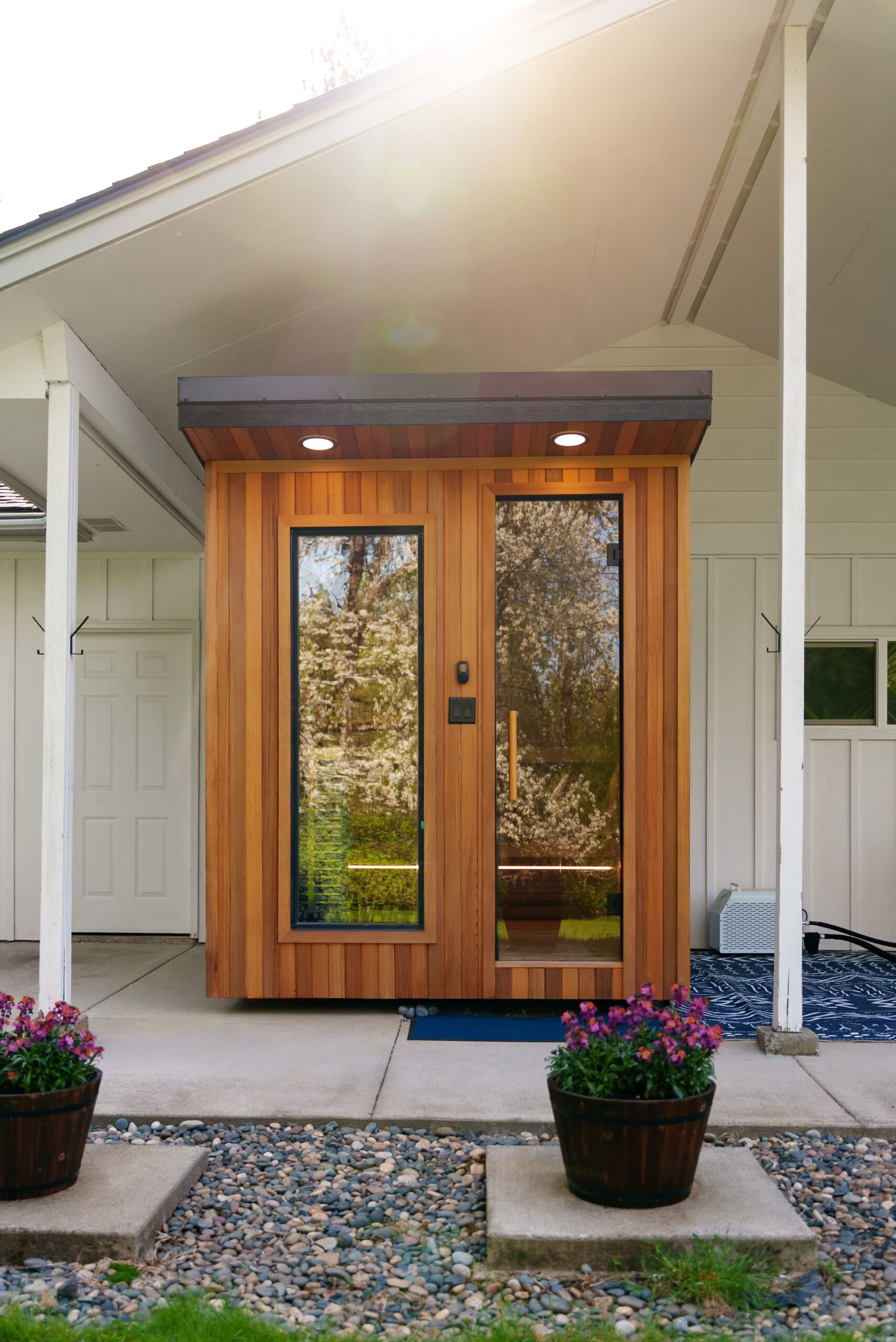 A modern wooden entryway with tall glass panels is attached to a white house. Two potted flowers sit on the stone path leading to the door, and sunlight shines through the trees in the background.