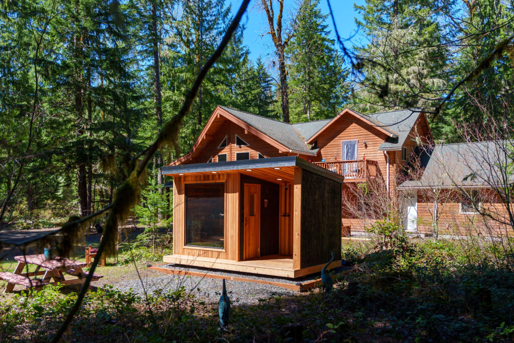 A small wooden cabin stands in front of a larger log house, surrounded by tall green trees. Sunlight filters through the forest, illuminating the cozy structures and a nearby picnic table.