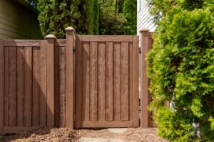 A brown wood-grain fence with a matching gate positioned between two evergreen bushes.